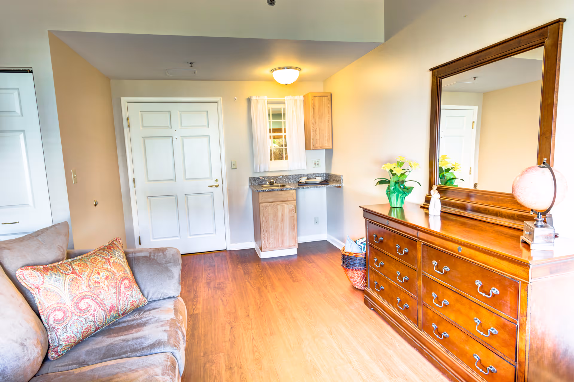 A cozy interior room featuring a brown cushioned sofa with a patterned pillow on the left, a wooden dresser with a large mirror on the right, and a small kitchenette area with a sink and cabinet in the center background. The room has wooden flooring and light-colored walls with a white door and window with sheer curtains.