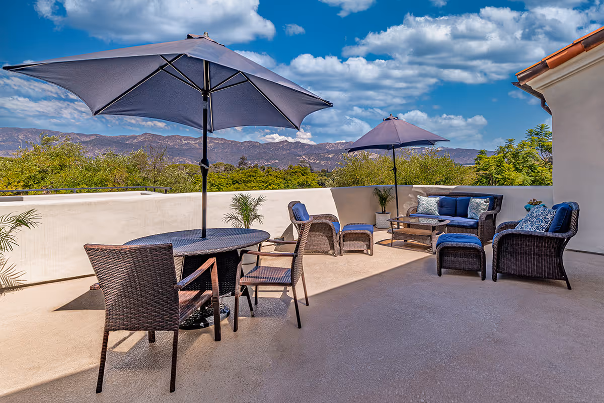 Outdoor patio area with wicker furniture including chairs, a loveseat, ottomans, and tables. Two large umbrellas provide shade. The patio overlooks greenery and distant mountains under a partly cloudy blue sky.
