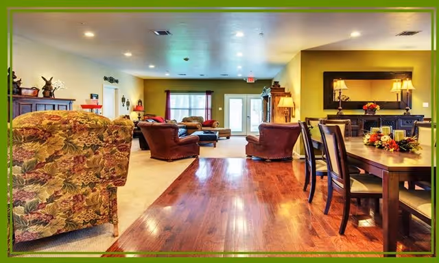 A spacious and well-lit living area in a senior living facility featuring a mix of floral and solid upholstered armchairs, a wooden dining table with chairs, a large mirror above a sideboard, and a window with curtains letting in natural light.