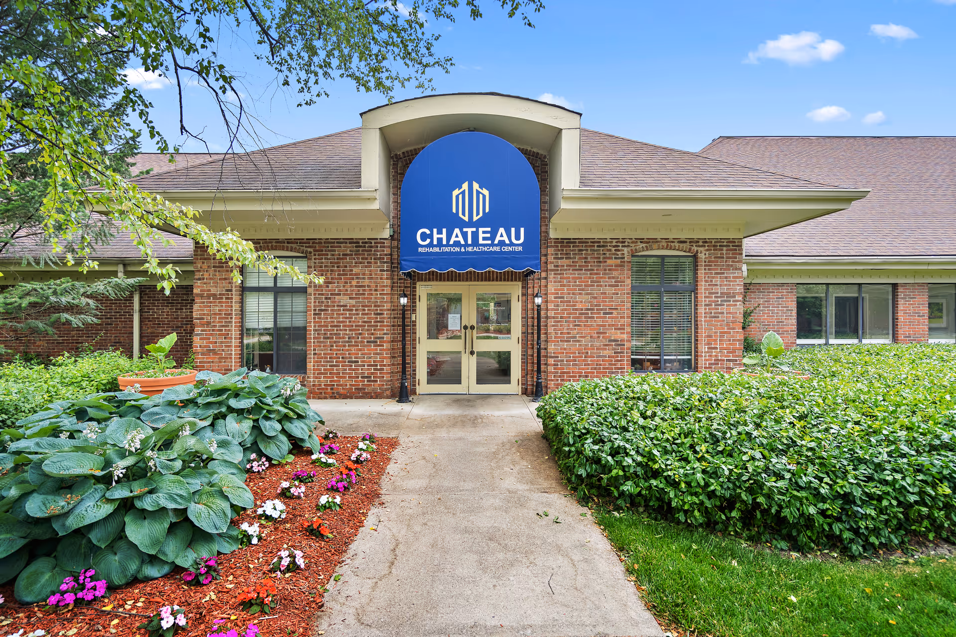 Front entrance of the Chateau Rehabilitation and Healthcare Center brick building with a blue awning, walkway, and landscaped flower beds.