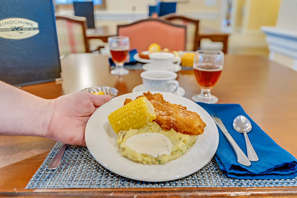 A plate of breaded fish, mashed potatoes with gravy and a corn on the cob being set on a dining table in a dining room.