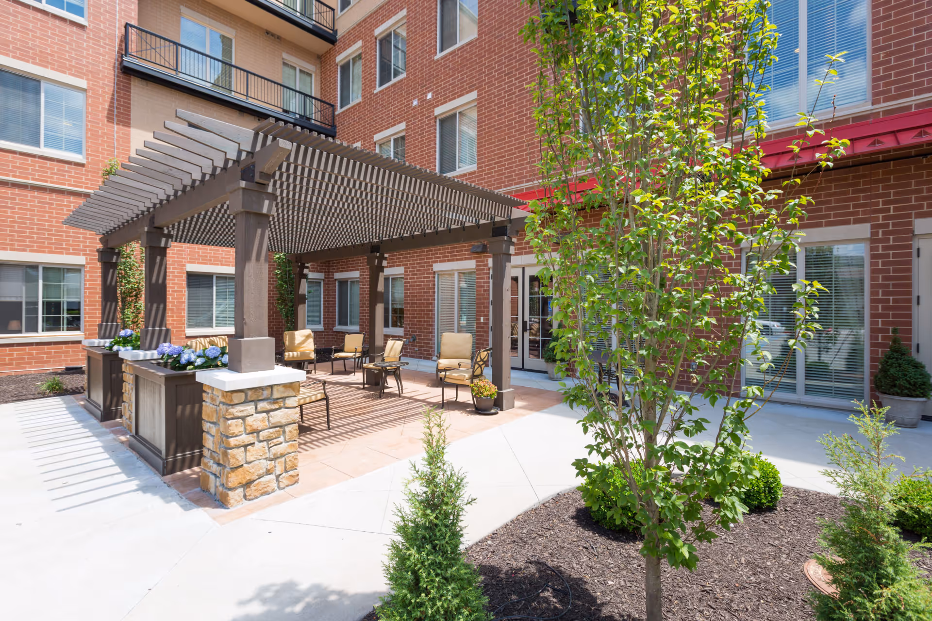 Sunlit outdoor courtyard featuring a wooden pergola with seating, planters, and a brick building facade.