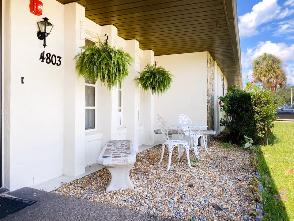 Outdoor seating area next to a white building with the number 4803 on the wall. The area features a stone bench, a white metal table with two matching chairs, and two hanging green ferns. There is a small garden with bushes and a palm tree in the background under a partly cloudy blue sky.