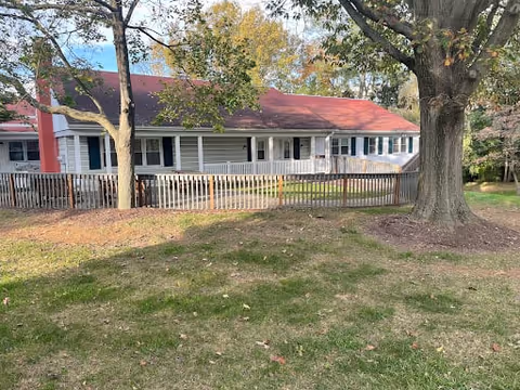 Single-story building with white siding and a red roof, surrounded by a wooden fence and trees with autumn foliage, viewed from a grassy area.
