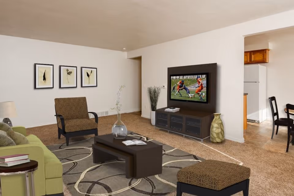 A cozy living room with a green sofa, a patterned armchair, and a matching ottoman. There is a dark wooden coffee table with a glass vase and an open book on it. On the wall, three framed botanical prints are displayed. A flat-screen TV mounted on a dark wooden entertainment unit shows a football game. The room has beige carpet and light-colored walls. In the background, a kitchen and dining area with a refrigerator and chairs are visible.