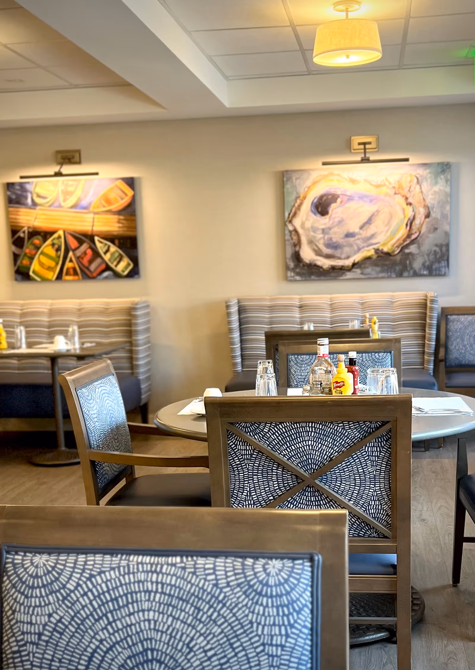 Interior view of a dining area with round tables and chairs featuring patterned backs. The tables are set with glasses, condiments, and napkins. The background shows striped cushioned bench seating and two colorful paintings on the wall, one depicting boats and the other an abstract design resembling an oyster shell.