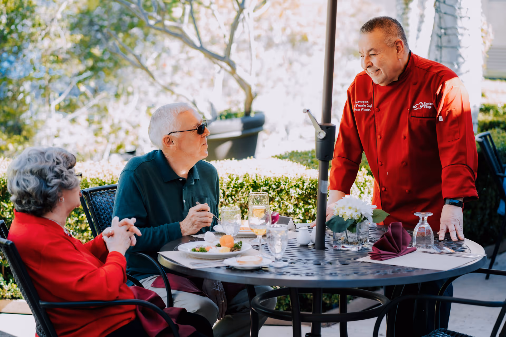 Two older adults seated at a round outdoor dining table being served by a chef in a red jacket.