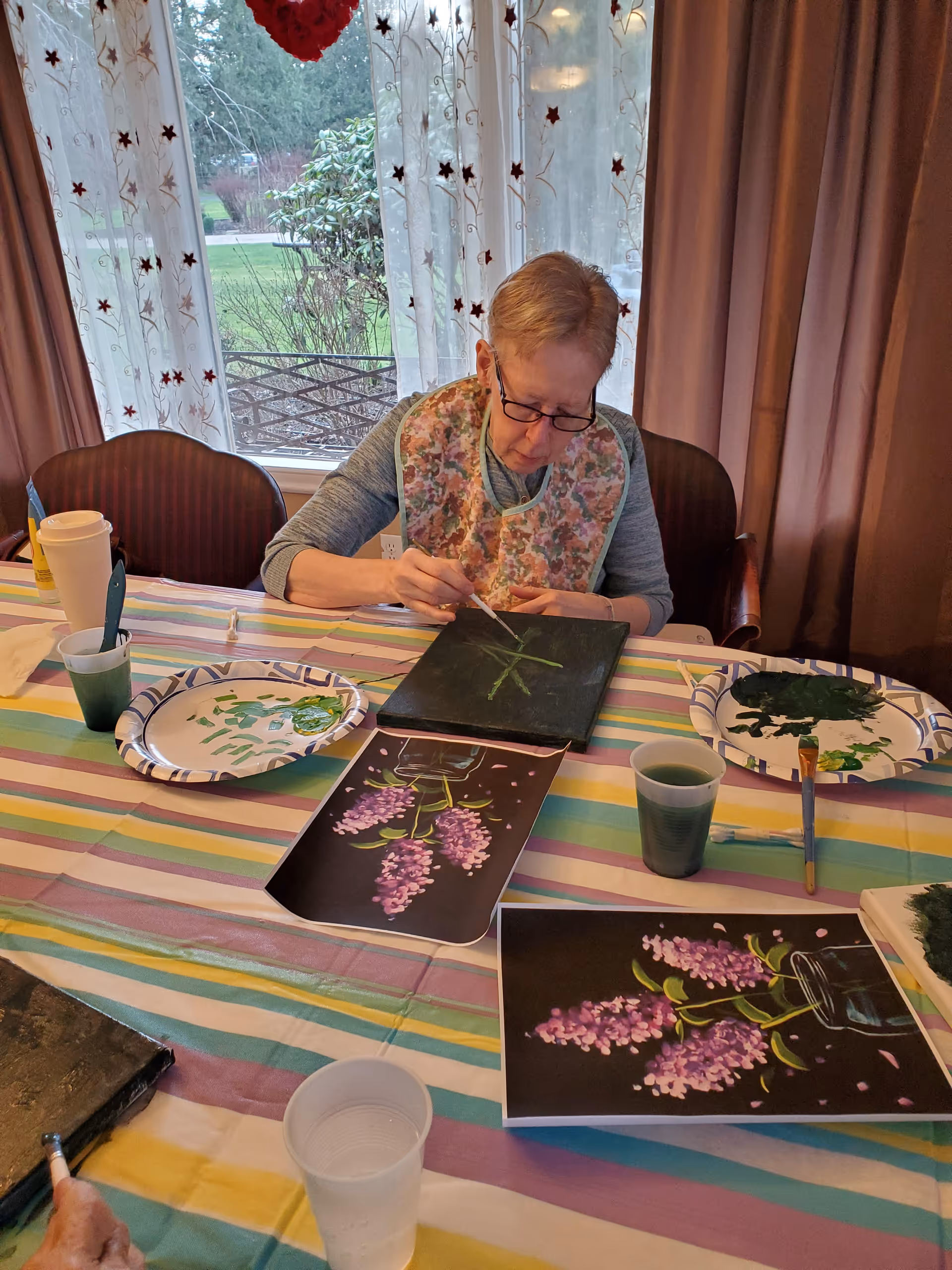 An elderly woman wearing glasses and a floral bib is seated at a table covered with a colorful striped tablecloth, painting on a small canvas. There are paint supplies, including brushes, cups with paint, and paper plates with paint, as well as printed images of purple flowers on the table. Behind her, sheer curtains with star patterns cover a window showing a green outdoor area.