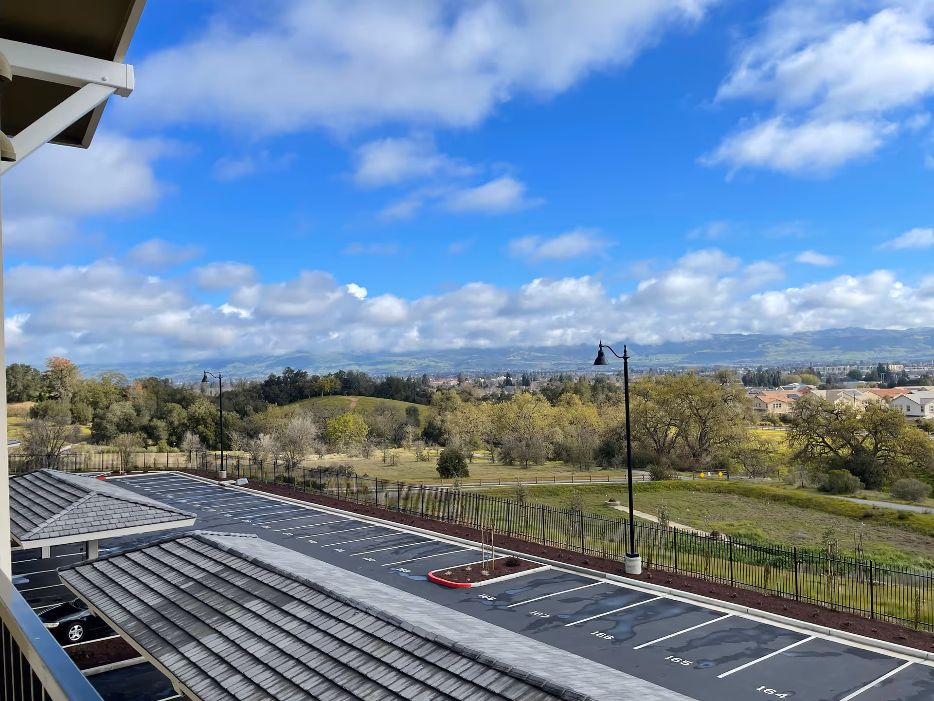 View from a balcony overlooking a parking lot with numbered parking spaces, a fence, grassy fields, trees, and hills in the distance under a partly cloudy blue sky.