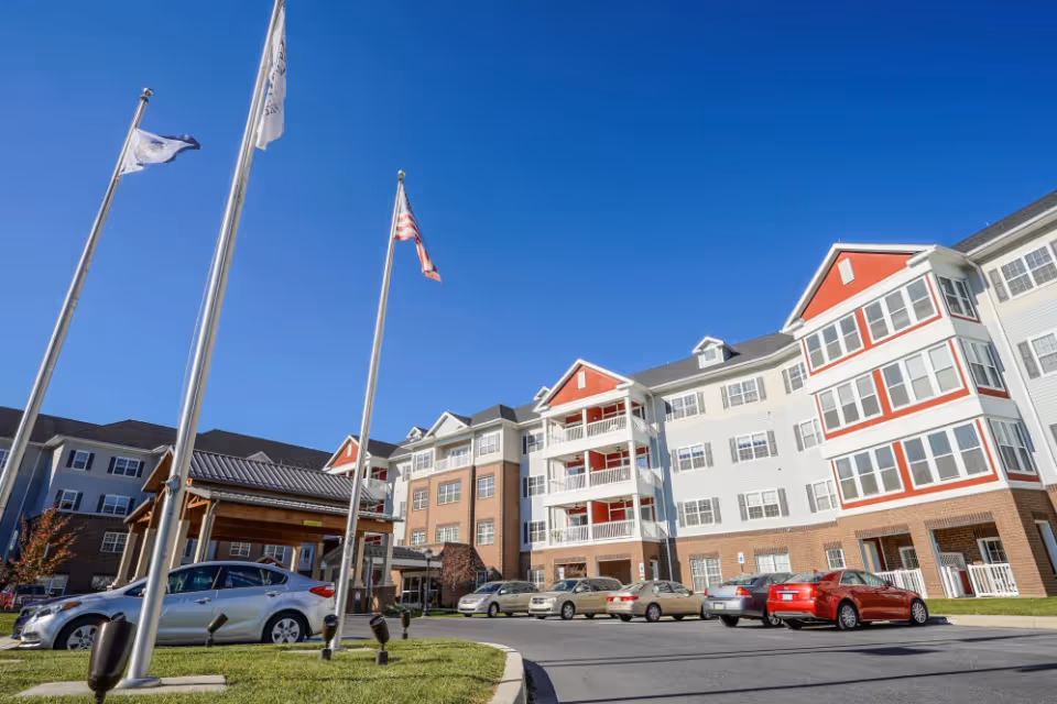 Exterior view of a multi-story senior living facility building with white and red accents, several parked cars in front, and three flagpoles with flags against a clear blue sky.
