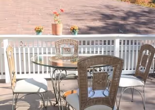 Outdoor patio area with a round glass table and four wicker chairs with white cushions. The patio has a white railing and three small potted plants placed on top of the railing. The background shows a roof and some greenery.