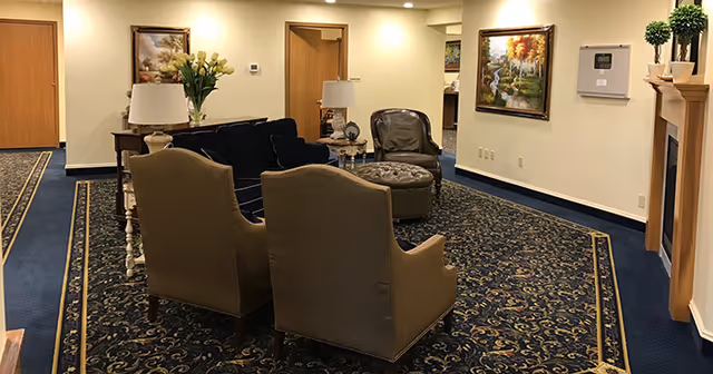 A cozy living room area with two beige armchairs, a black sofa, a round ottoman, and two table lamps on side tables. The room has a dark patterned carpet with a blue border, cream-colored walls, framed paintings, and a fireplace with plants on the mantel.