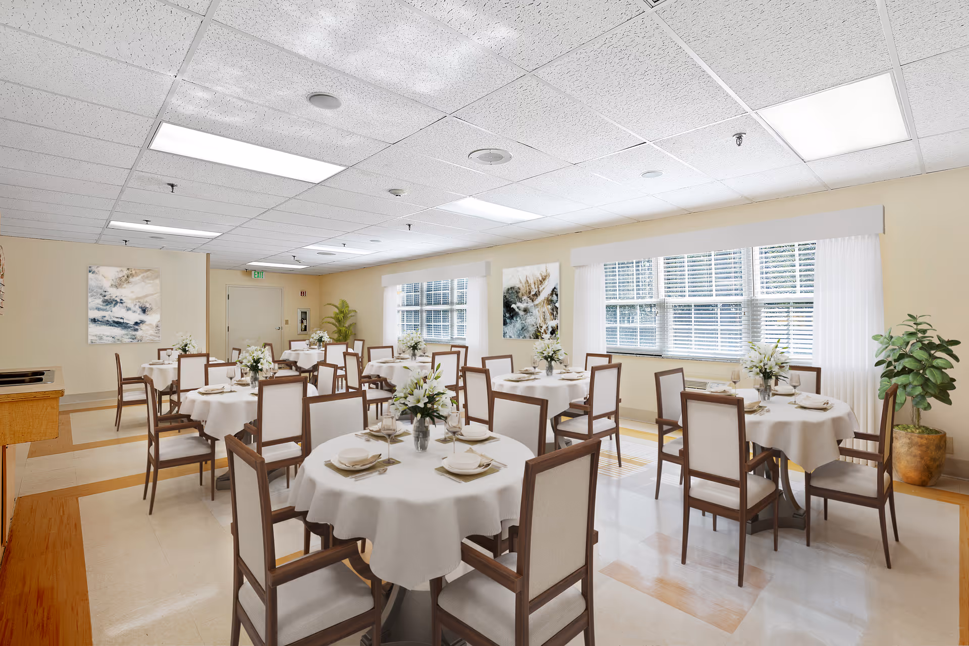 A bright dining room with several round tables covered with white tablecloths, each set with plates, napkins, and floral centerpieces. The room has large windows with white blinds, beige walls adorned with abstract paintings, and wooden chairs with cushioned seats arranged around the tables. There are also potted plants in the corners, and the ceiling has recessed lighting.
