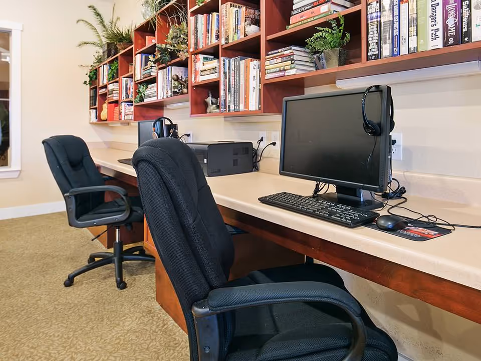 A computer workstation area with two black office chairs and two desktop computers on a long beige countertop. Above the countertop are wooden shelves filled with books and decorative plants. The room has beige walls and carpeted flooring.