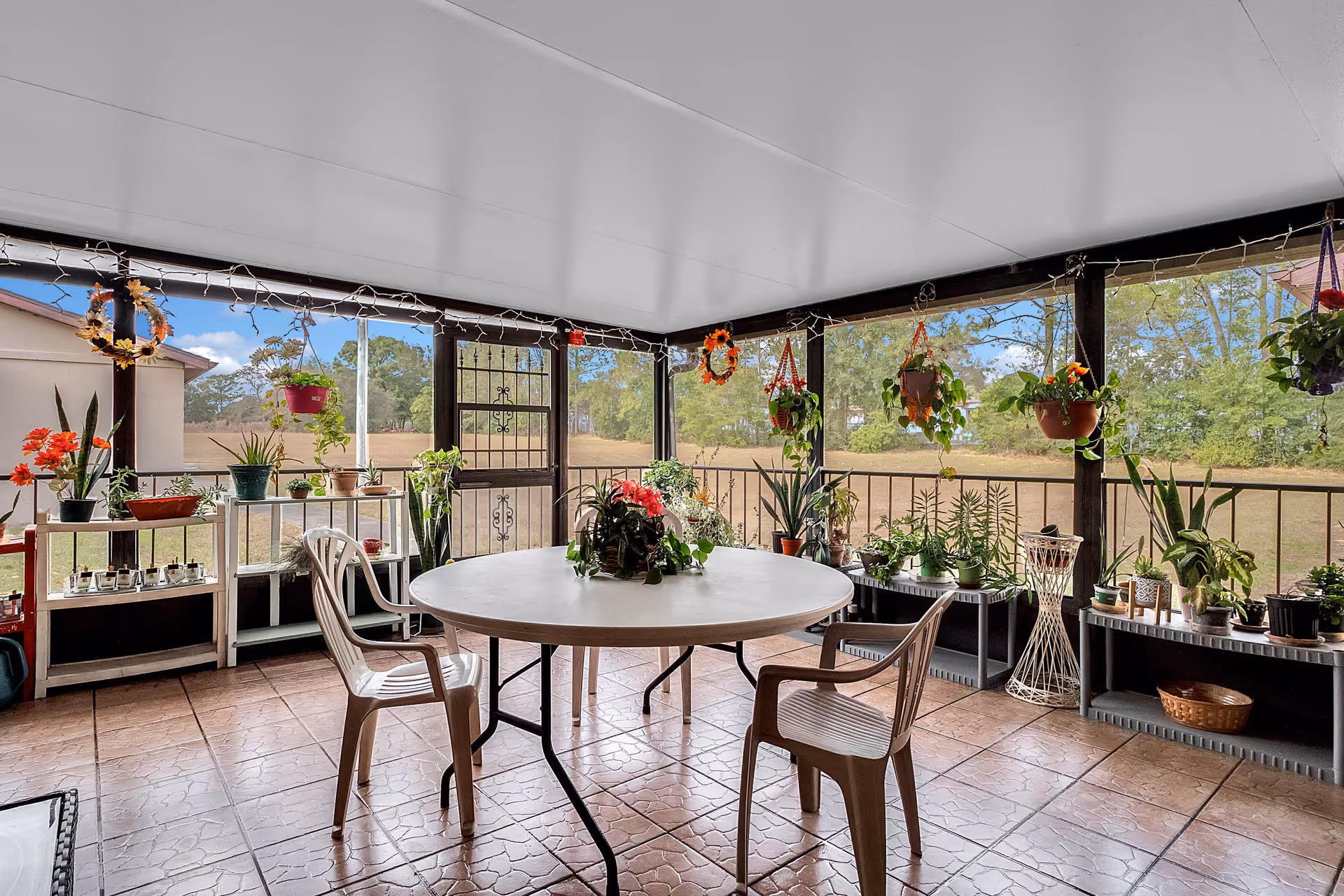 A covered patio area with a round white table and two plastic chairs. The patio is decorated with numerous potted plants on shelves and hanging from the ceiling. The area is enclosed with screened windows, providing a view of a grassy field and trees outside. The floor is tiled with a textured pattern.