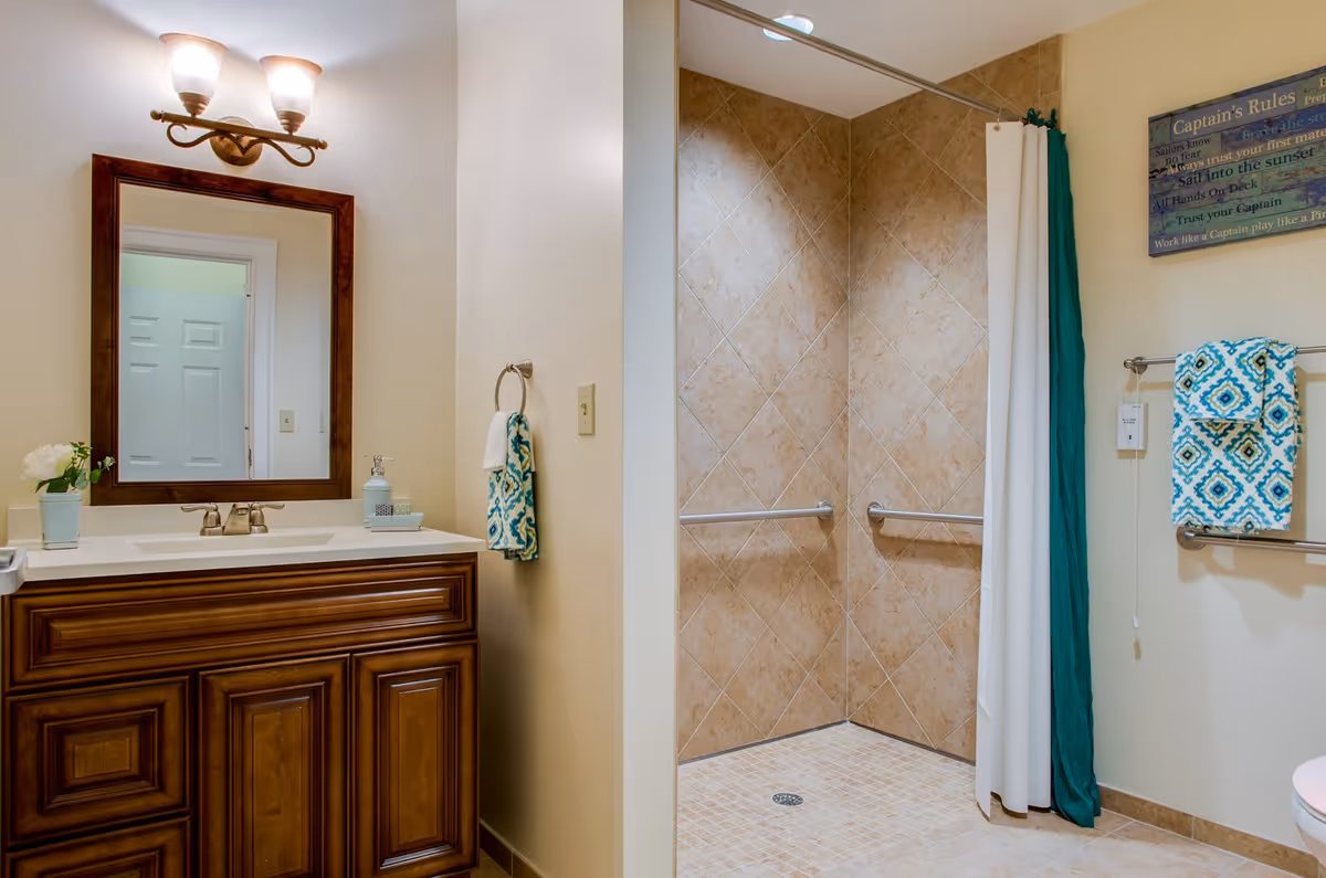 A bathroom with a wooden vanity featuring a white countertop and a mirror above it. There is a light fixture with three bulbs above the mirror. To the right, there is a walk-in shower with beige tiled walls and floor, equipped with metal grab bars and a white shower curtain with a green accent. A towel rack with a patterned blue and white towel is mounted on the wall next to the shower, along with a decorative sign above it.