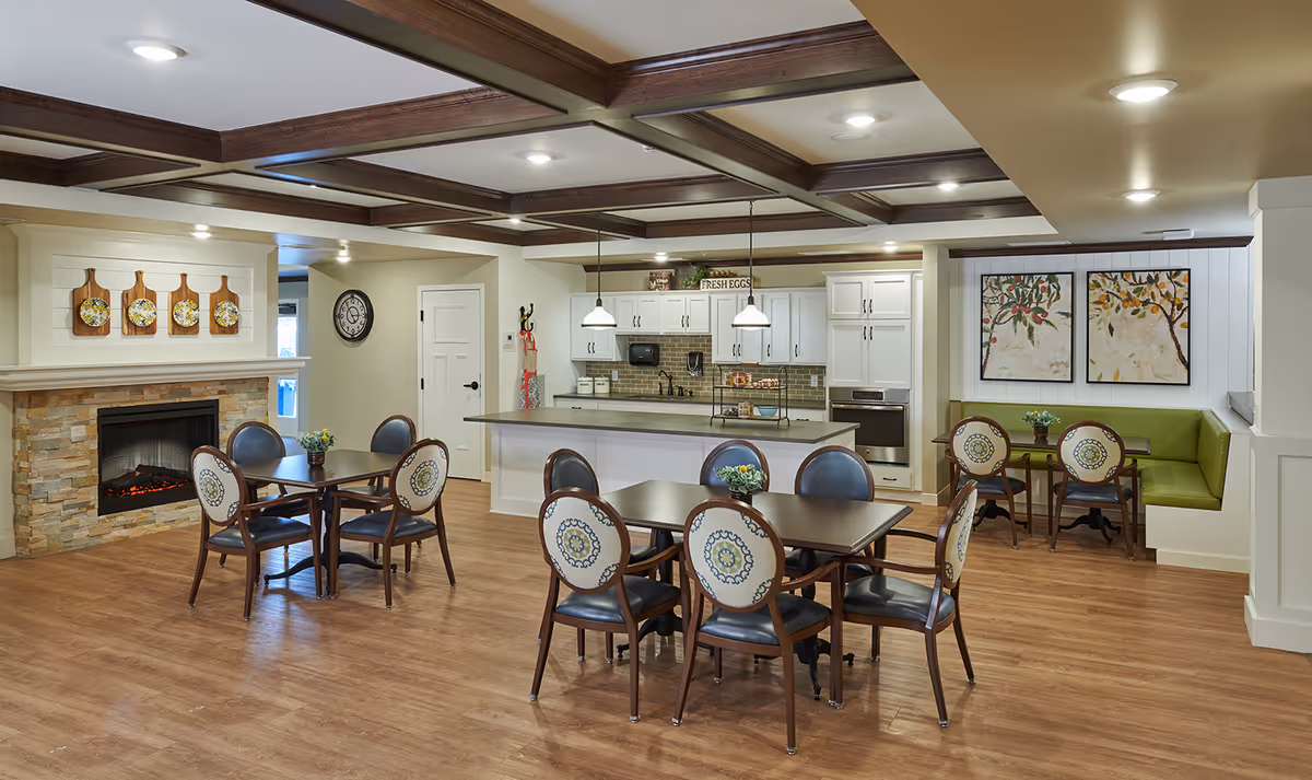 A cozy dining area in a senior living facility featuring wooden tables and chairs with patterned upholstery. The room has a modern kitchen with white cabinets, a stone fireplace, and decorative wall art. The ceiling has wooden beams and recessed lighting, and there is a green cushioned bench seating area in the corner.
