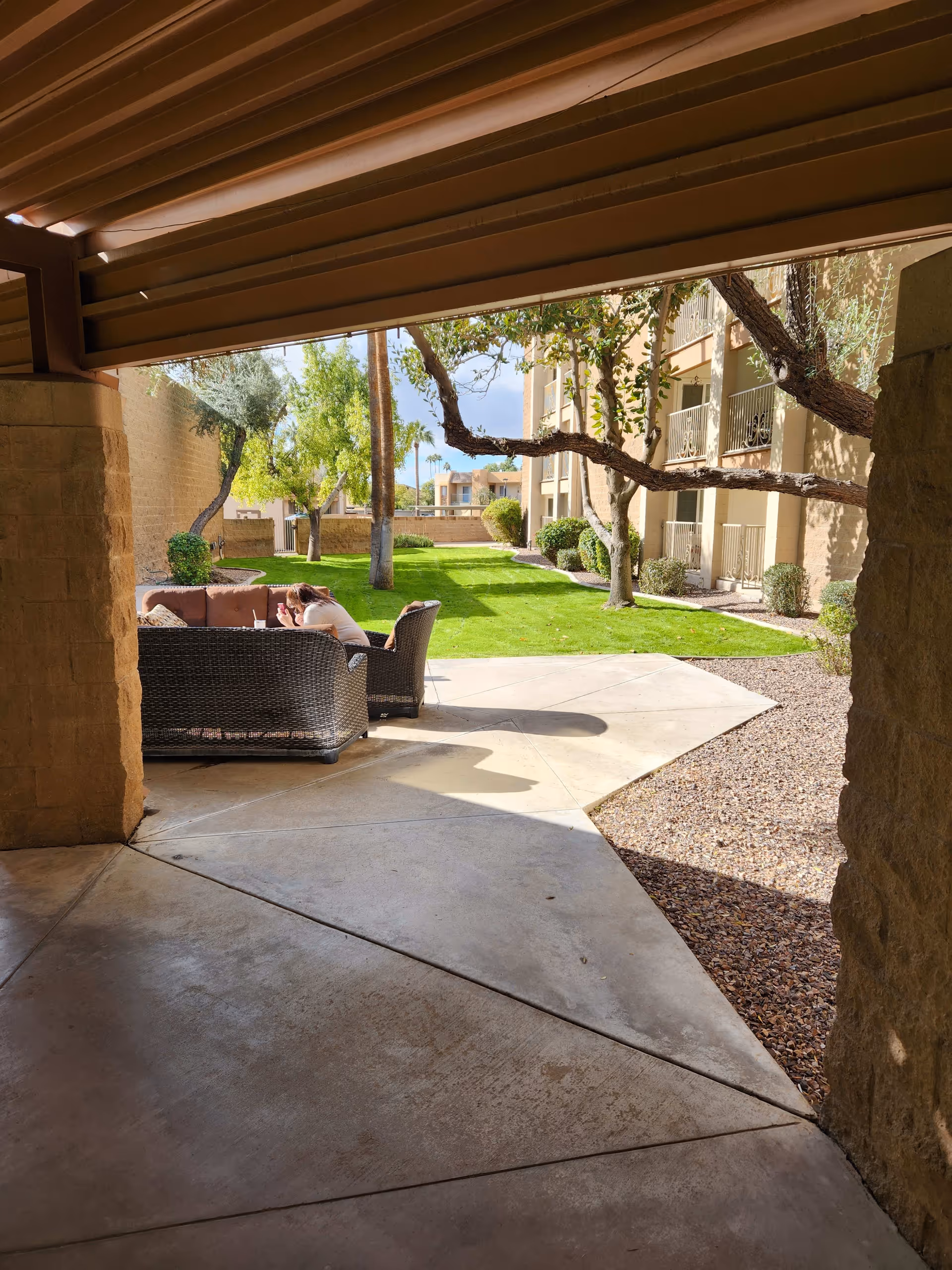 View from a shaded patio area looking out onto a green lawn with trees and shrubs. There is outdoor wicker furniture with cushions and a person sitting on one of the chairs. In the background, there are multi-story residential buildings with balconies.