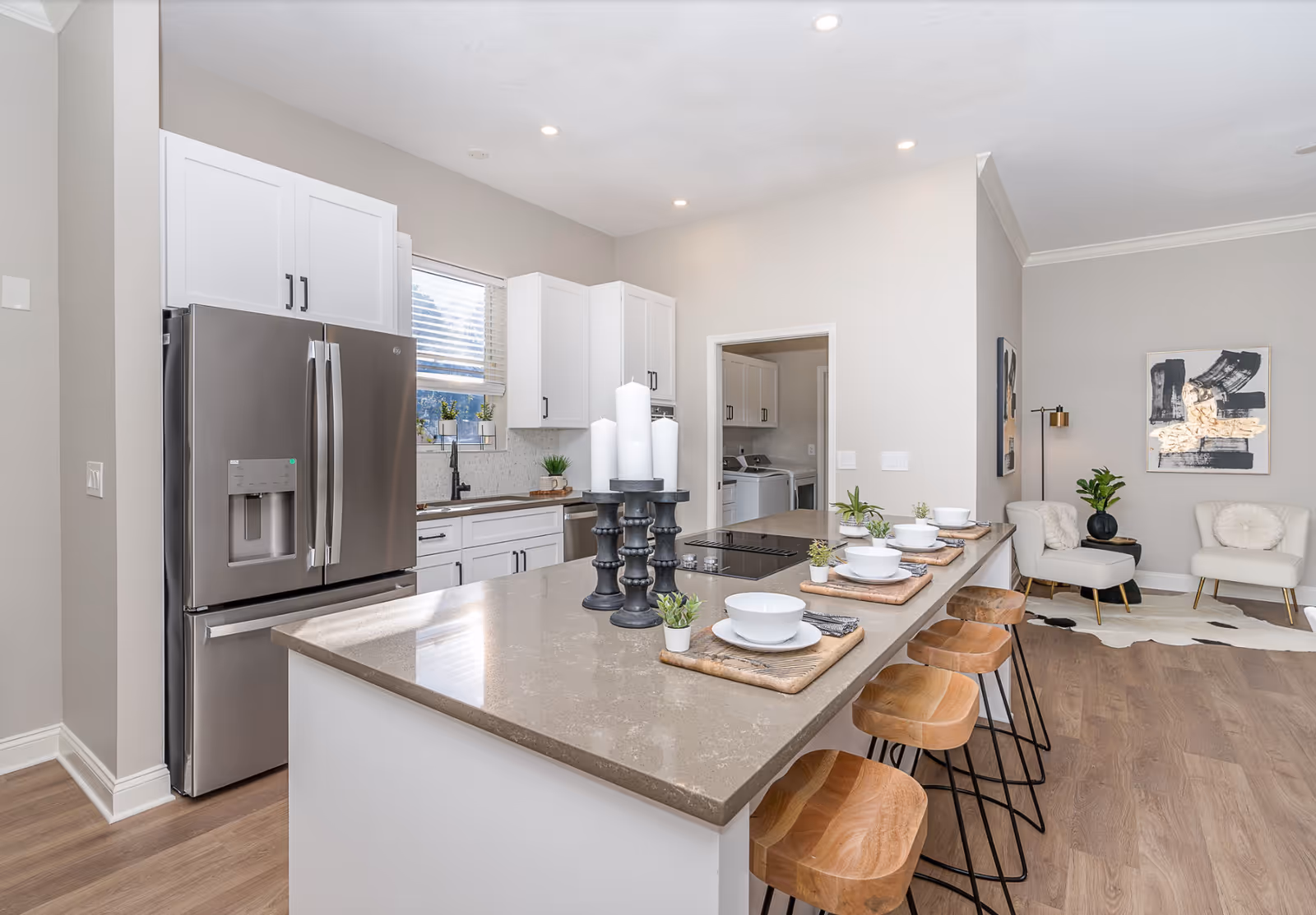 Modern kitchen with a large island countertop featuring four wooden bar stools, white bowls and plates set on wooden placemats, and decorative candles. Stainless steel refrigerator and white cabinetry with a window above the sink. Adjacent living area with two white chairs, a black side table, a floor lamp, and abstract wall art.