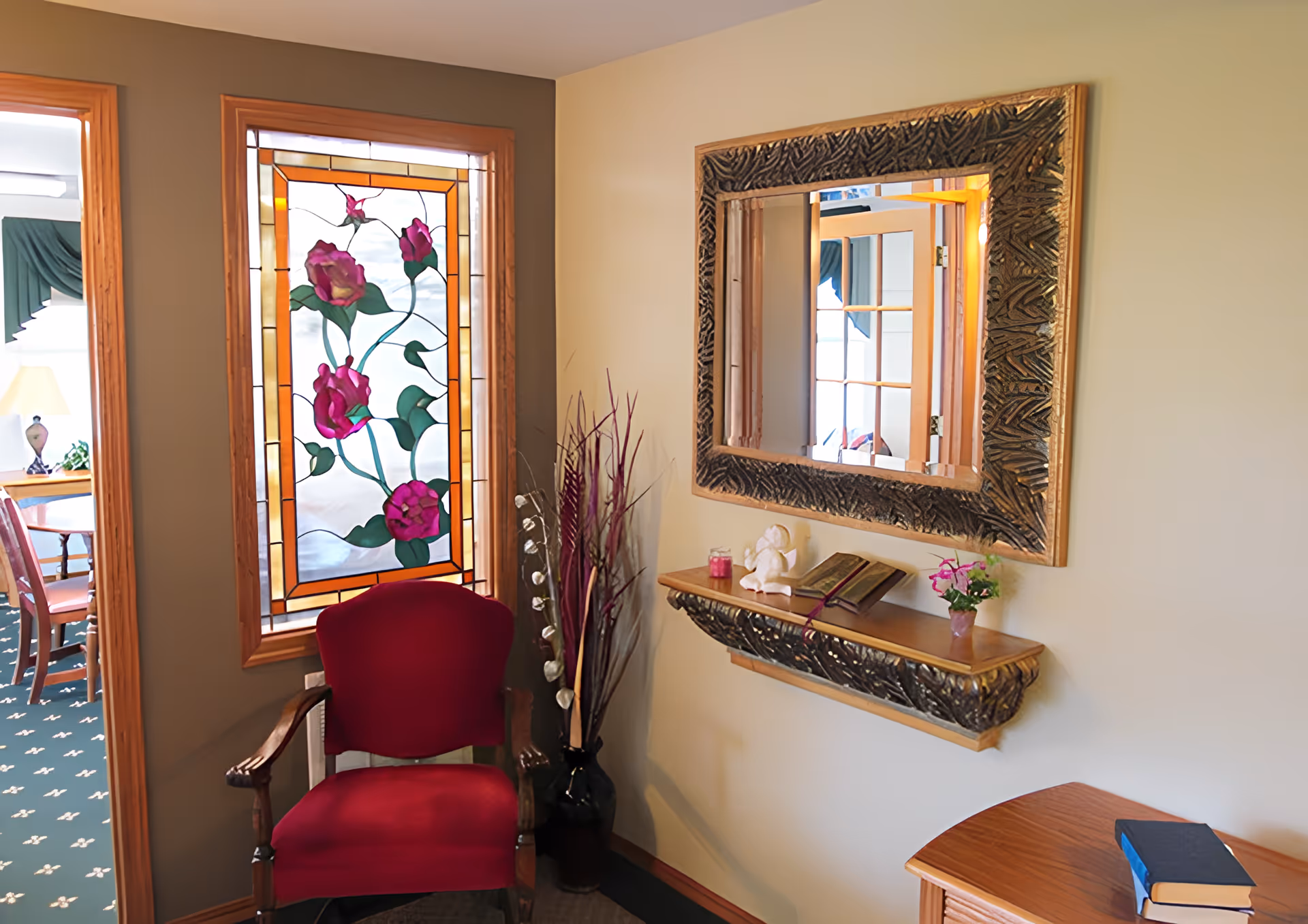 A cozy corner of an interior room featuring a red upholstered wooden armchair, a decorative stained glass window with pink flowers, a wall-mounted shelf with a small angel figurine, an open book, a candle, and a small potted plant. Above the shelf is a large ornate mirror reflecting part of the room. A wooden table with a closed book is partially visible in the foreground.