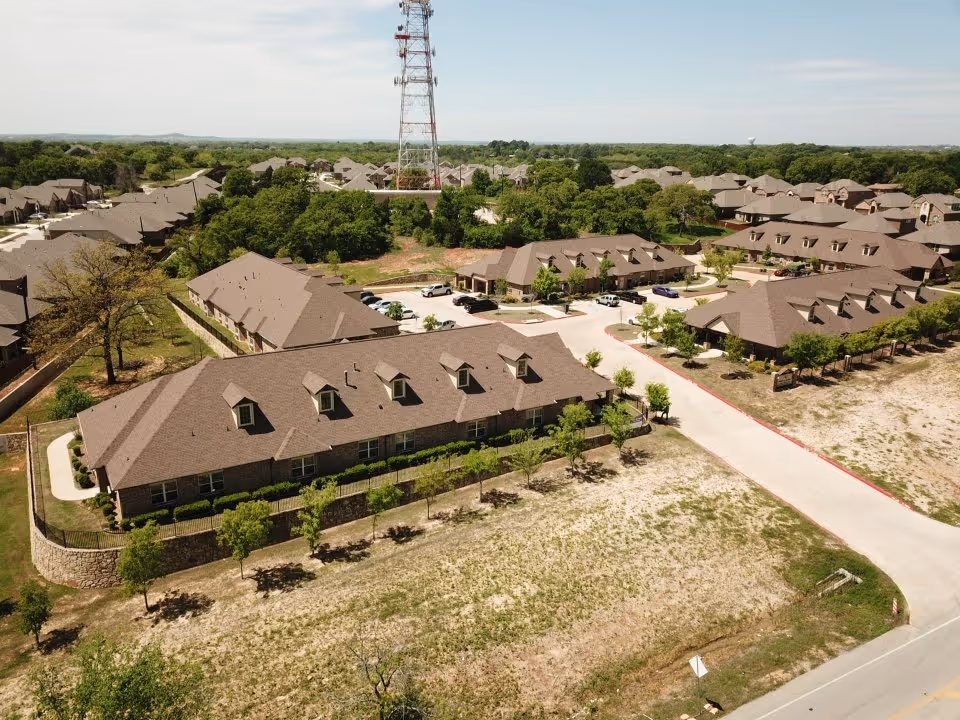 Aerial view of Mayberry Gardens Assisted Living and Memory Care Homes showing multiple single-story buildings with brown roofs, surrounded by trees and greenery. A paved road runs through the complex, and several cars are parked near the buildings. A tall communication tower is visible in the background.