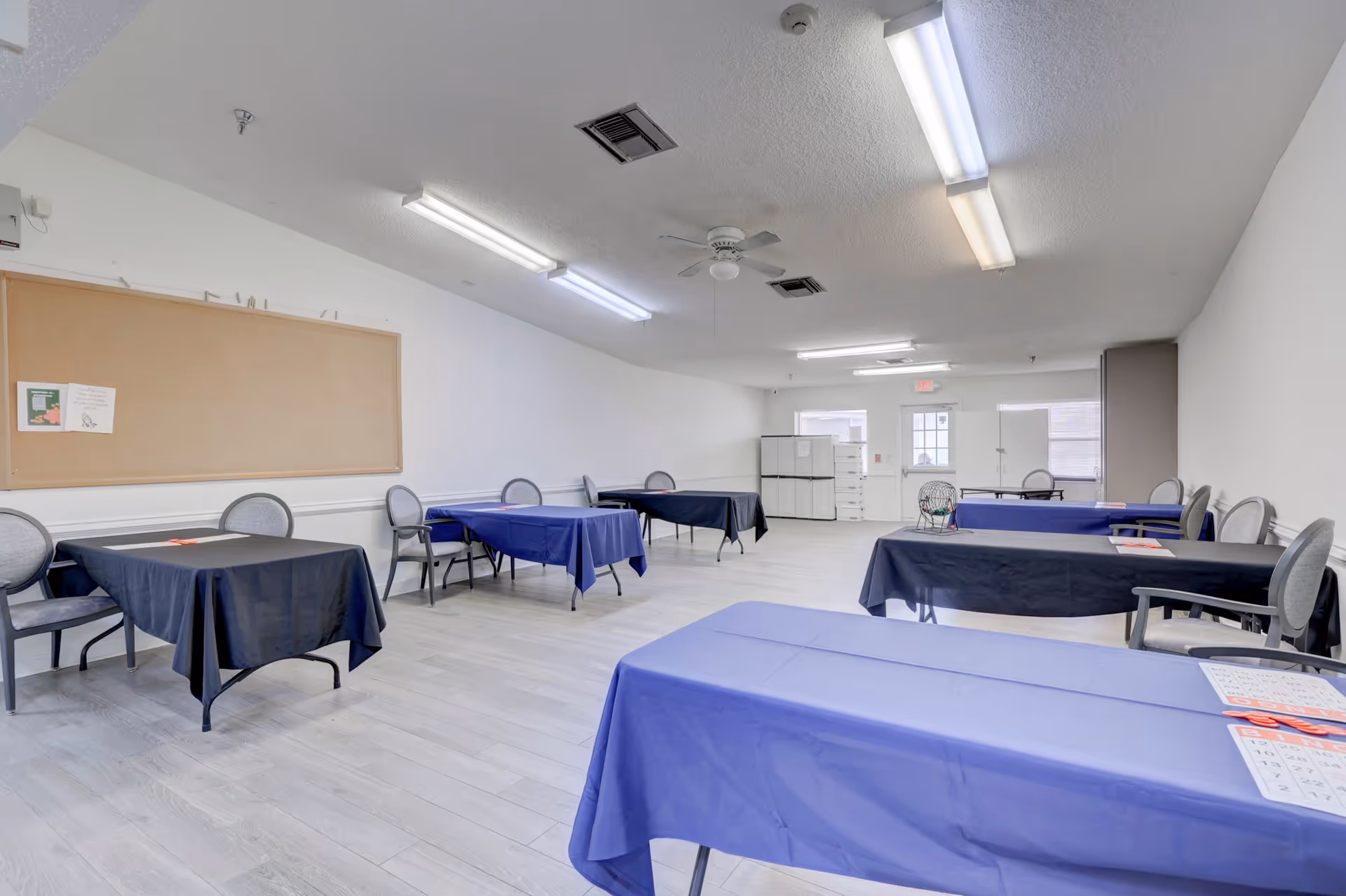 A spacious room with several tables covered in dark blue tablecloths and surrounded by chairs. The room has light-colored flooring, white walls, fluorescent ceiling lights, a ceiling fan, and a large bulletin board on one wall. There are some papers and a bingo cage on the tables, suggesting it may be used for activities or dining.