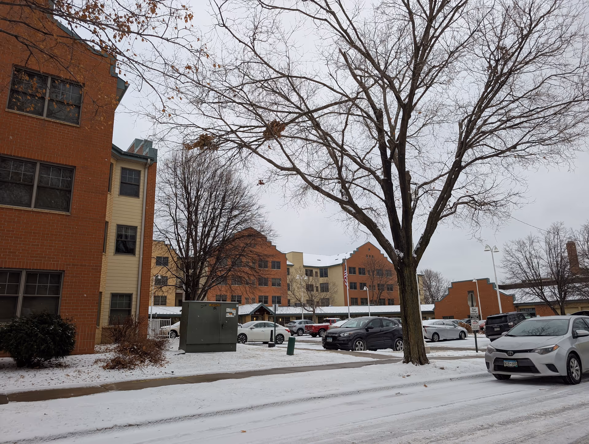Snow-covered parking lot with several parked cars in front of a multi-story brick and beige building. Leafless trees are visible, and the sky is overcast. A sign near the parking lot reads 'River Village East Catholic Elder Care.'