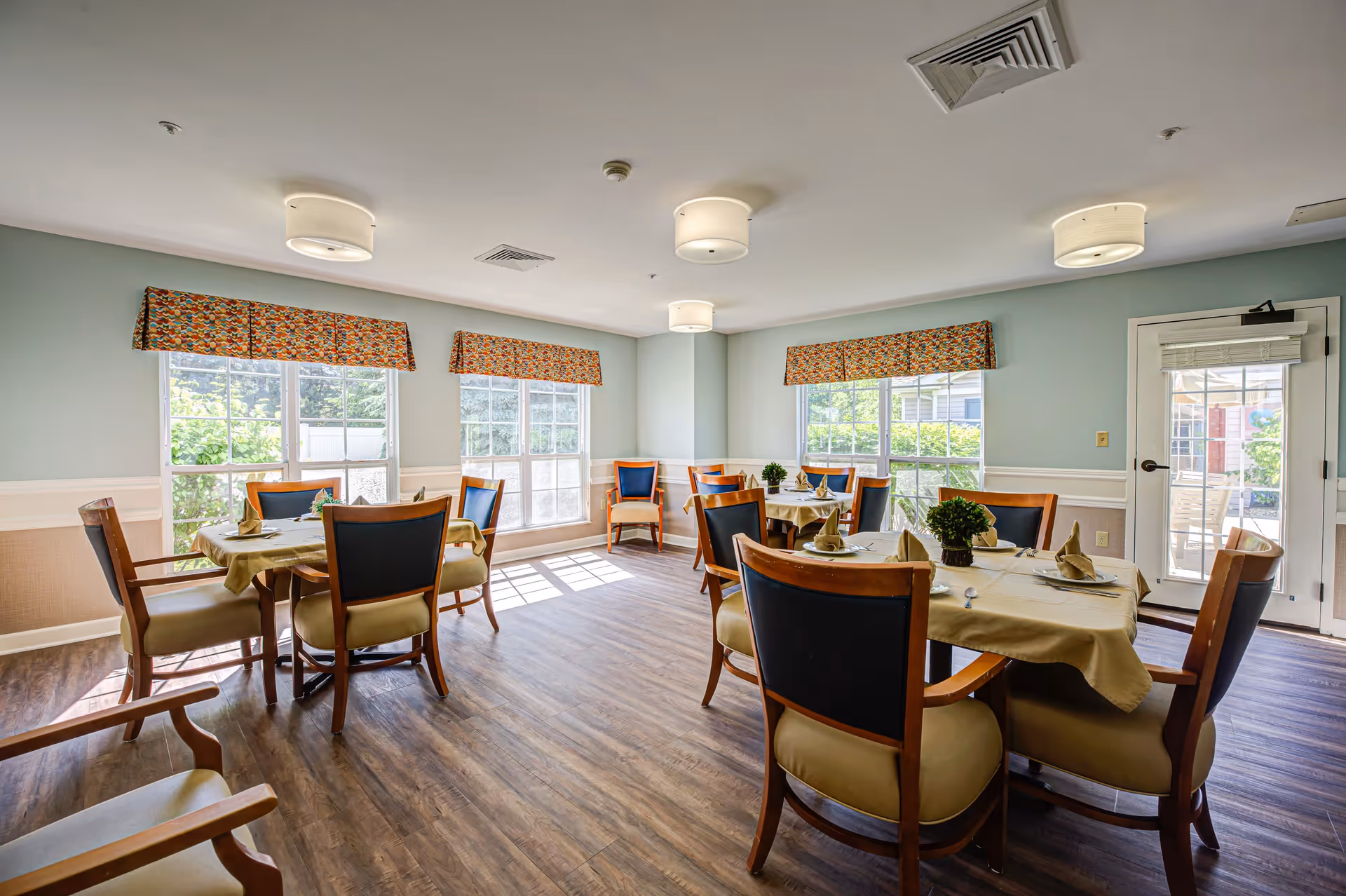 A bright dining room with several round tables covered with beige tablecloths, each set with folded napkins, cutlery, and small green plants as centerpieces. The room has large windows with colorful valances, light blue walls with white wainscoting, wood flooring, and ceiling lights. There is a door leading outside on the right side.
