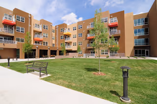 Sunlit courtyard with a grassy lawn, benches and lamp posts surrounded by a multi-story apartment building with balconies.