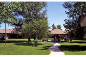 A sunny outdoor area at Devonshire Care Center featuring a paved walkway, green grass, several trees, and a building with a brown roof in the background.