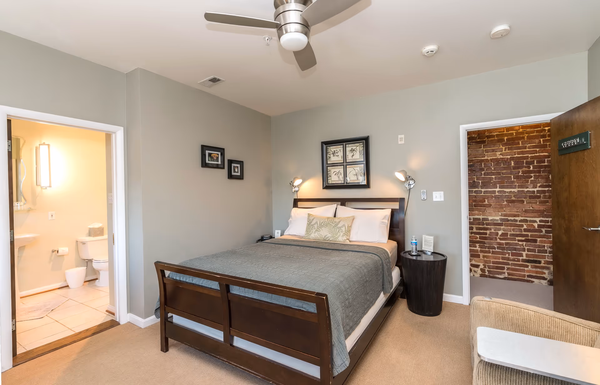 A cozy bedroom with a wooden bed frame, gray bedding, and white pillows. The room has light gray walls with framed artwork above the bed and two wall-mounted reading lamps. To the left, there is an open door leading to a bathroom with a toilet and sink visible. To the right, there is another open door showing a brick wall in the adjacent room. A ceiling fan is mounted on the ceiling, and a small round bedside table with a water bottle and other items is next to the bed.