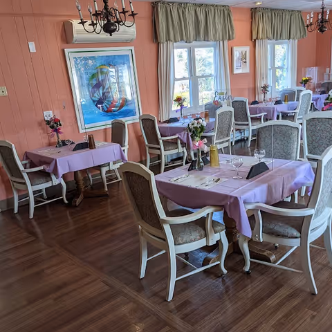Dining room with multiple tables covered in lavender tablecloths, set with place settings and floral centerpieces, surrounded by upholstered chairs.