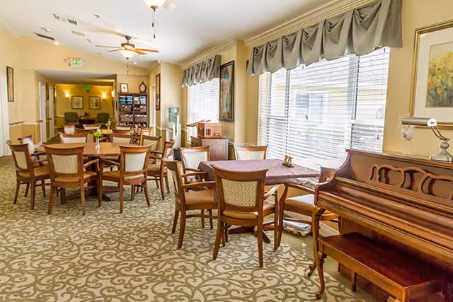 Bright communal dining/activity room with several round tables and chairs, a piano, and large windows letting in sunlight.