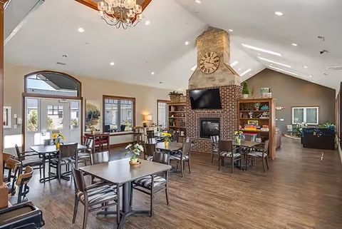 A spacious common area with multiple tables and chairs arranged on a wooden floor. There is a large brick fireplace with a clock above it and a mounted TV. The room has high ceilings with skylights and large windows letting in natural light. Shelves with plants and decorative items flank the fireplace. The entrance door and additional seating areas are visible in the background.