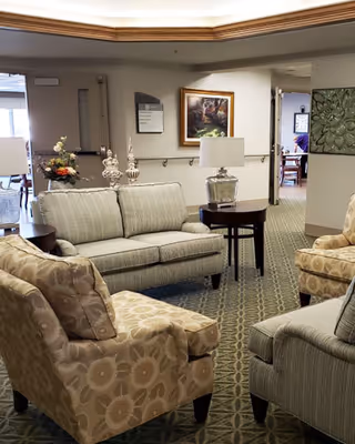 A cozy seating area in a senior living facility with a striped sofa, two patterned armchairs, a round wooden side table with a lamp, decorative vases, and framed artwork on the walls. The carpet has a geometric pattern and there is a hallway leading to another room in the background.