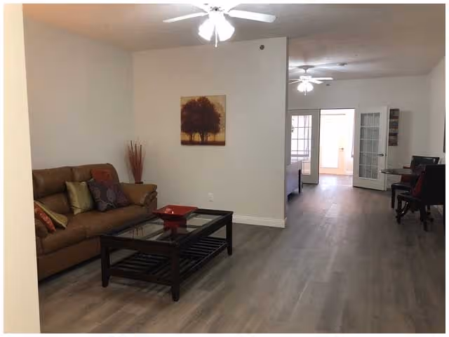 Interior view of a living room area in an assisted living facility with a brown leather sofa adorned with colorful pillows, a dark wooden coffee table with a red decorative tray, a ceiling fan with lights, and a painting of a tree on the wall. The room extends into a hallway with another ceiling fan and a glimpse of additional rooms and French doors.