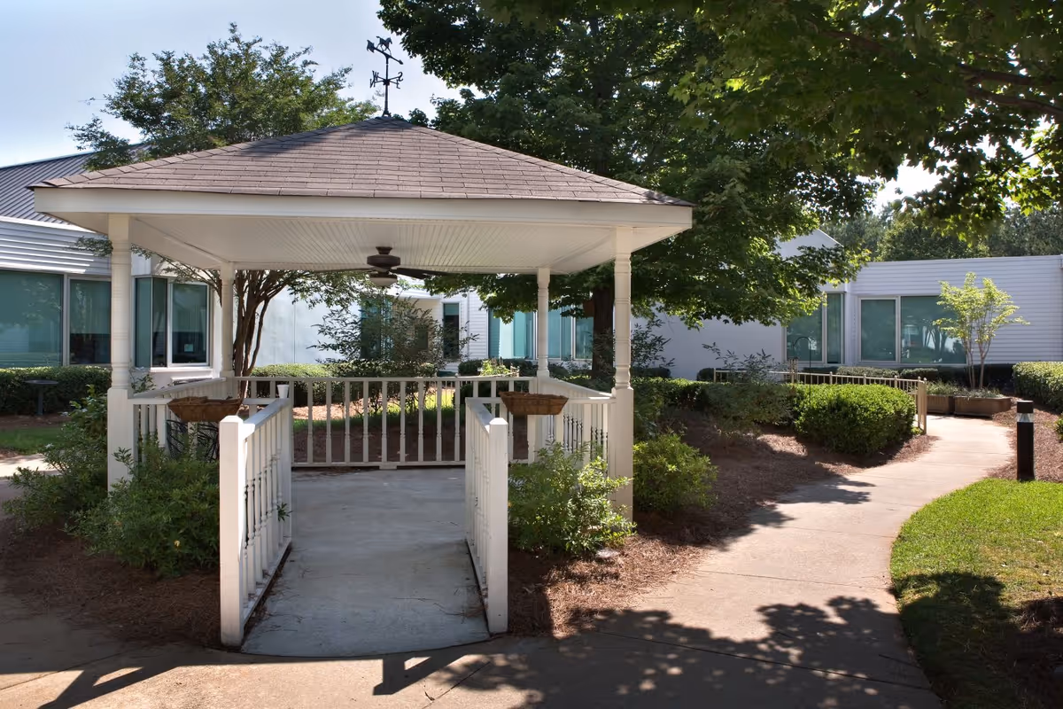 A white gazebo with a weather vane on top, surrounded by greenery and bushes, located in an outdoor garden area of a senior living facility. There is a concrete pathway leading to and from the gazebo, with trees and shrubs around, and the building with large windows visible in the background.