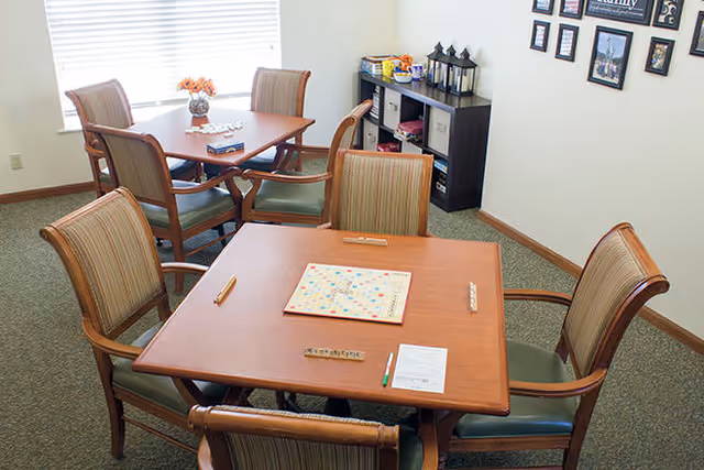 A well-lit room with two wooden tables, each surrounded by four wooden chairs with striped cushions. One table has a Scrabble game set up with letter tiles and racks, along with a pencil and a score sheet. The other table has a vase with flowers and some items on it. A black shelving unit against the wall holds various decorative items and games. Several framed pictures and quotes are hung on the wall.