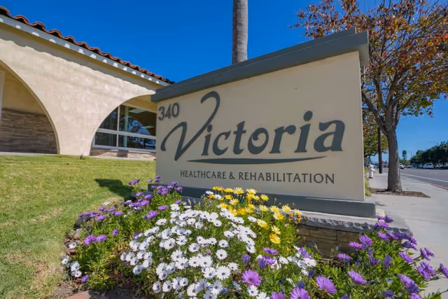 Entrance sign reading "Victoria Healthcare & Rehabilitation" with the number 340, set among flowering plants in front of the facility building and sidewalk.
