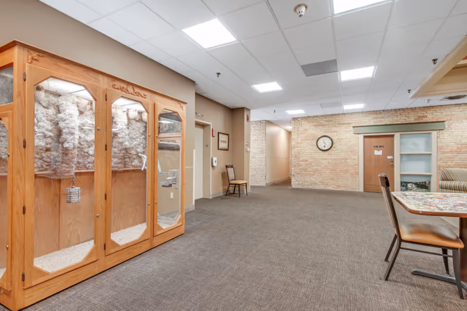 Interior view of a senior living facility hallway with a wooden display cabinet on the left, a chair against the wall, a clock on a brick wall, and a table with chairs on the right side.