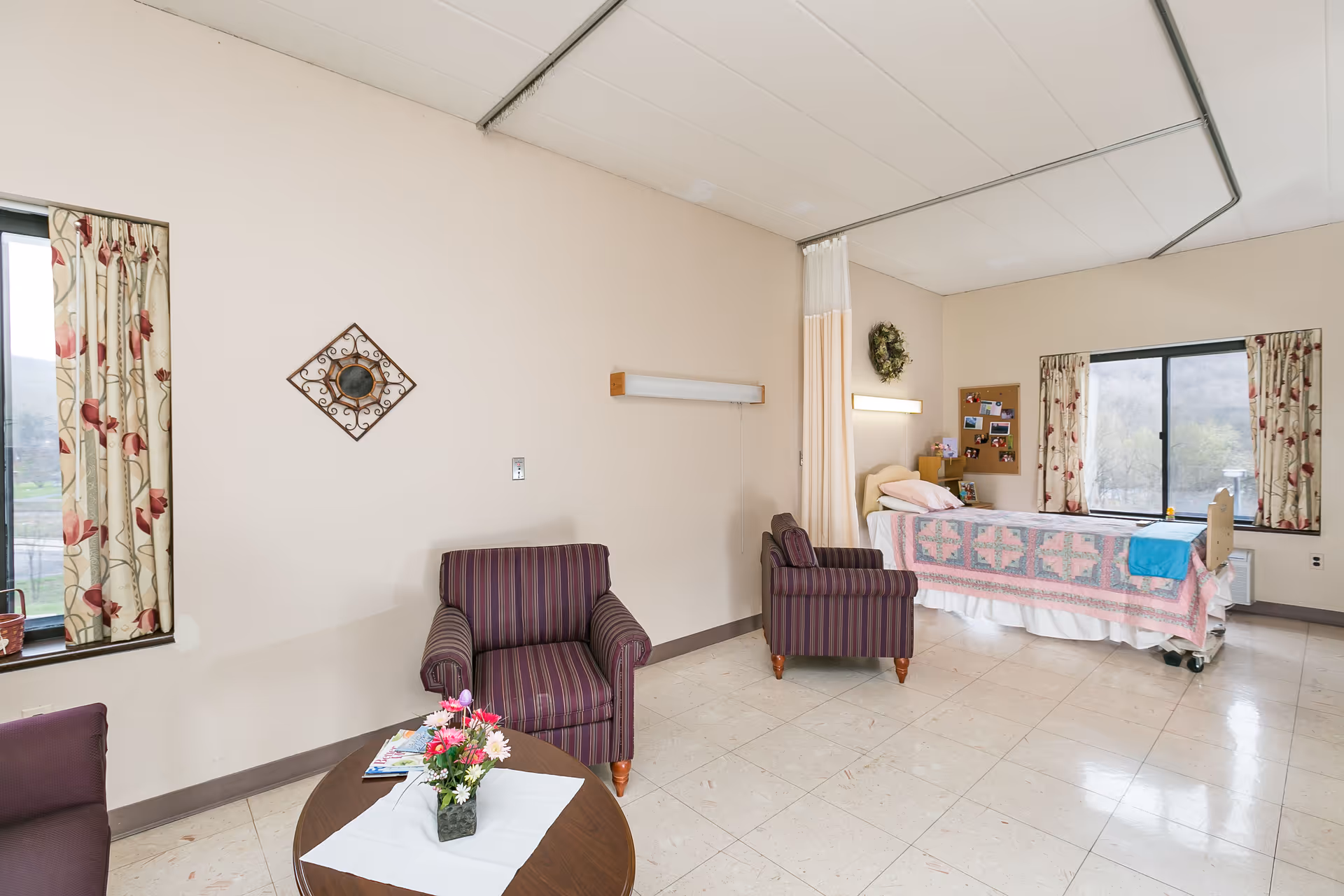 A nursing facility room with a hospital bed covered in a pink and blue quilt near a window with floral curtains. Two striped armchairs and a round table with a flower vase and magazines are in the foreground. The room has beige walls, tiled floor, and a privacy curtain rail on the ceiling.