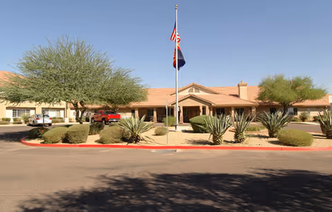 Exterior view of a single-story building with a tiled roof, desert landscaping including cacti and bushes, and an American flag and another flag on a flagpole in front of the building under a clear blue sky.