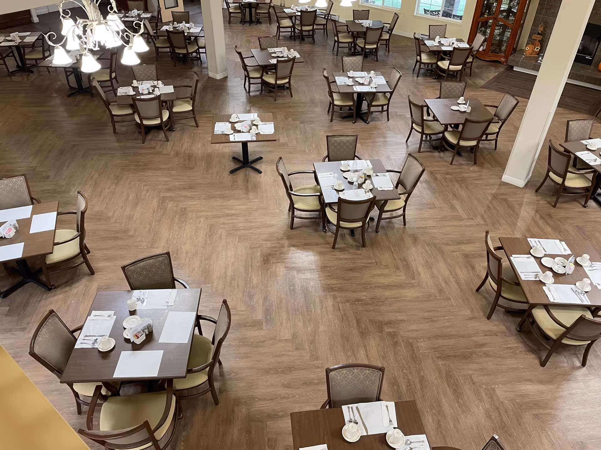 A spacious dining room with multiple square wooden tables, each set with white placemats, cups, saucers, and utensils. The room has wooden flooring and is well-lit with a chandelier hanging from the ceiling. Chairs with cushioned seats surround each table, and large windows allow natural light to enter the space.