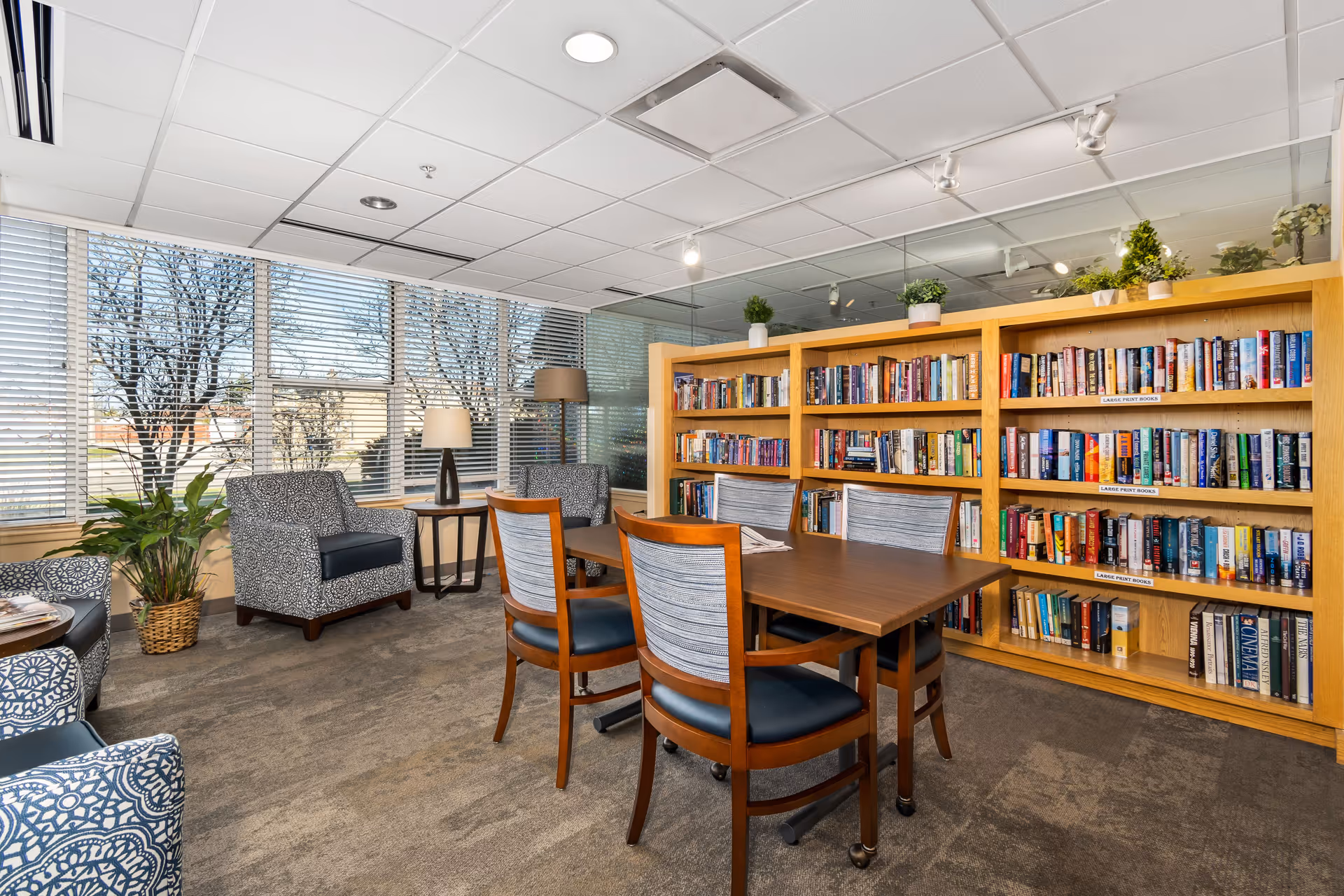 A cozy reading room with a wooden table surrounded by four chairs with blue cushions. Behind the table is a large wooden bookshelf filled with books. The room has large windows with blinds, two patterned armchairs, a side table with a lamp, a floor lamp, and several potted plants. The ceiling has recessed lighting and a white grid pattern.