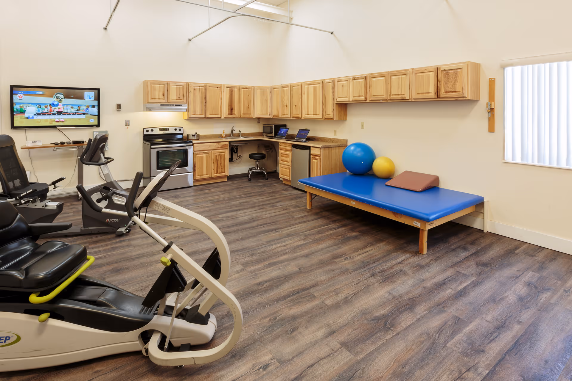 Interior therapy/fitness room with exercise machines, a treatment table with exercise balls, and a kitchenette along the back wall.