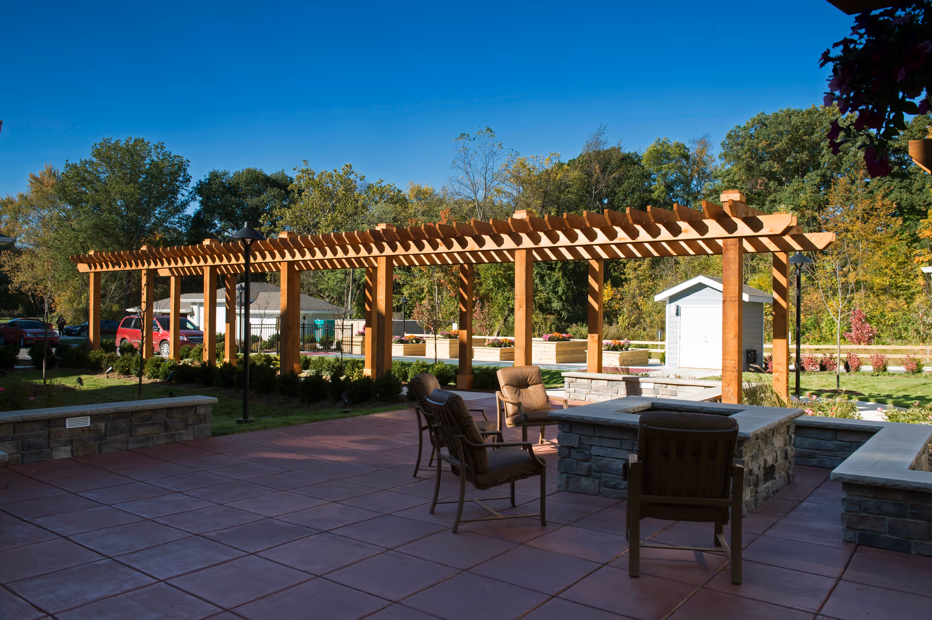 Outdoor patio area with cushioned chairs arranged around a stone fire pit. A wooden pergola structure extends across the background with greenery and trees beyond. A small white shed and parked cars are visible in the distance under a clear blue sky.