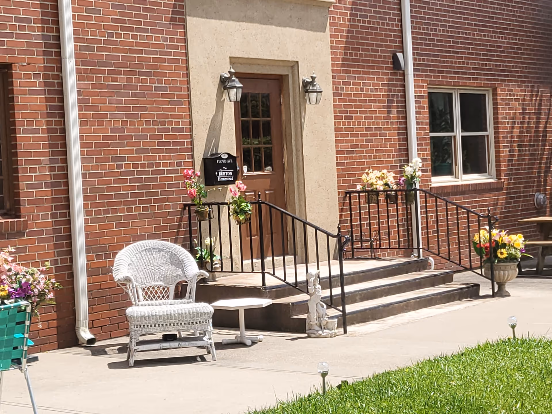Outdoor entrance area of The New Burton Homestead with a brown door framed by a beige stone surround, two wall-mounted lantern lights, and a black metal railing decorated with flower pots. A white wicker chair and a small white table are placed on the concrete patio near the entrance. The building is made of red brick, and there is a green lawn in the foreground.
