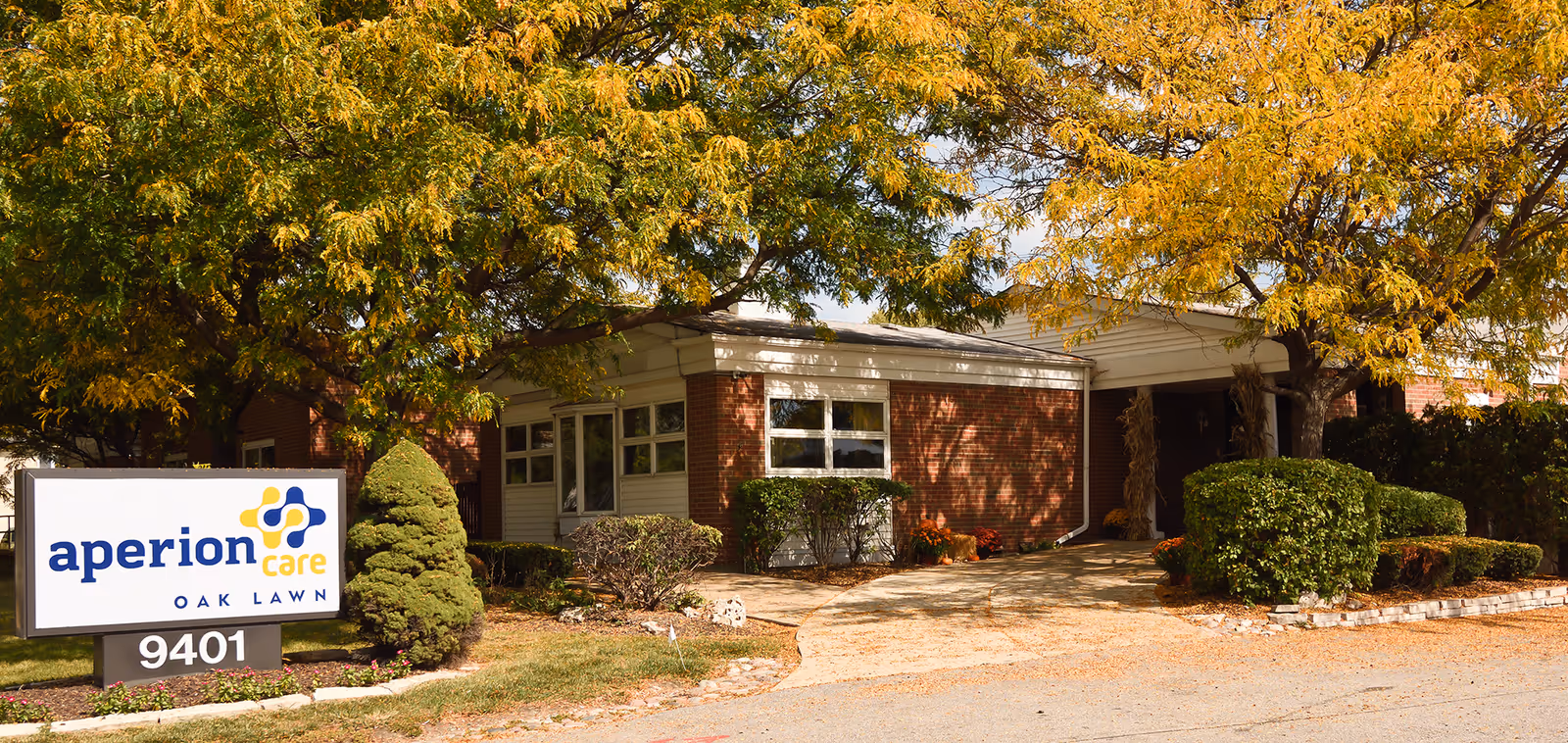 Front entrance of Aperion Care Oak Lawn with a brick facade, driveway, landscaped shrubs and a sign reading "aperion care OAK LAWN".