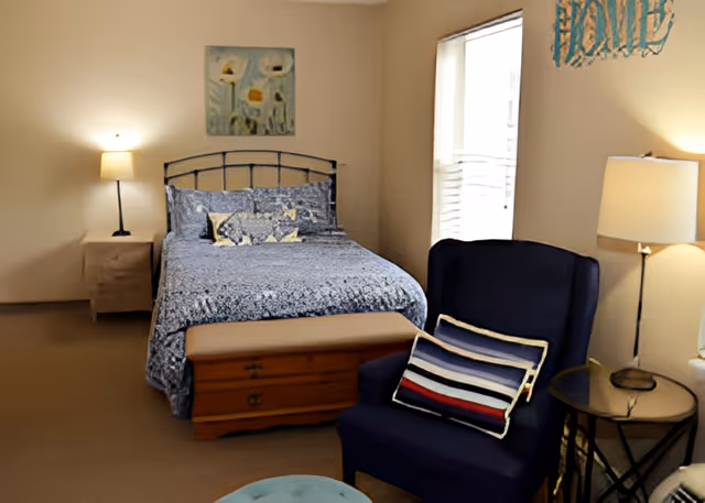 A cozy bedroom with a bed covered in a blue patterned bedspread, a wooden chest at the foot of the bed, a navy blue armchair with a striped pillow, two bedside tables each with a lamp, a window with blinds, and wall decorations including a painting and a sign that says 'HOME'.