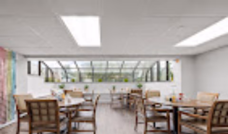 A bright dining room with multiple wooden tables and chairs arranged neatly. The room features large windows and a glass ceiling section allowing natural light to fill the space. Small plants are placed on the windowsills, and the walls are painted white.